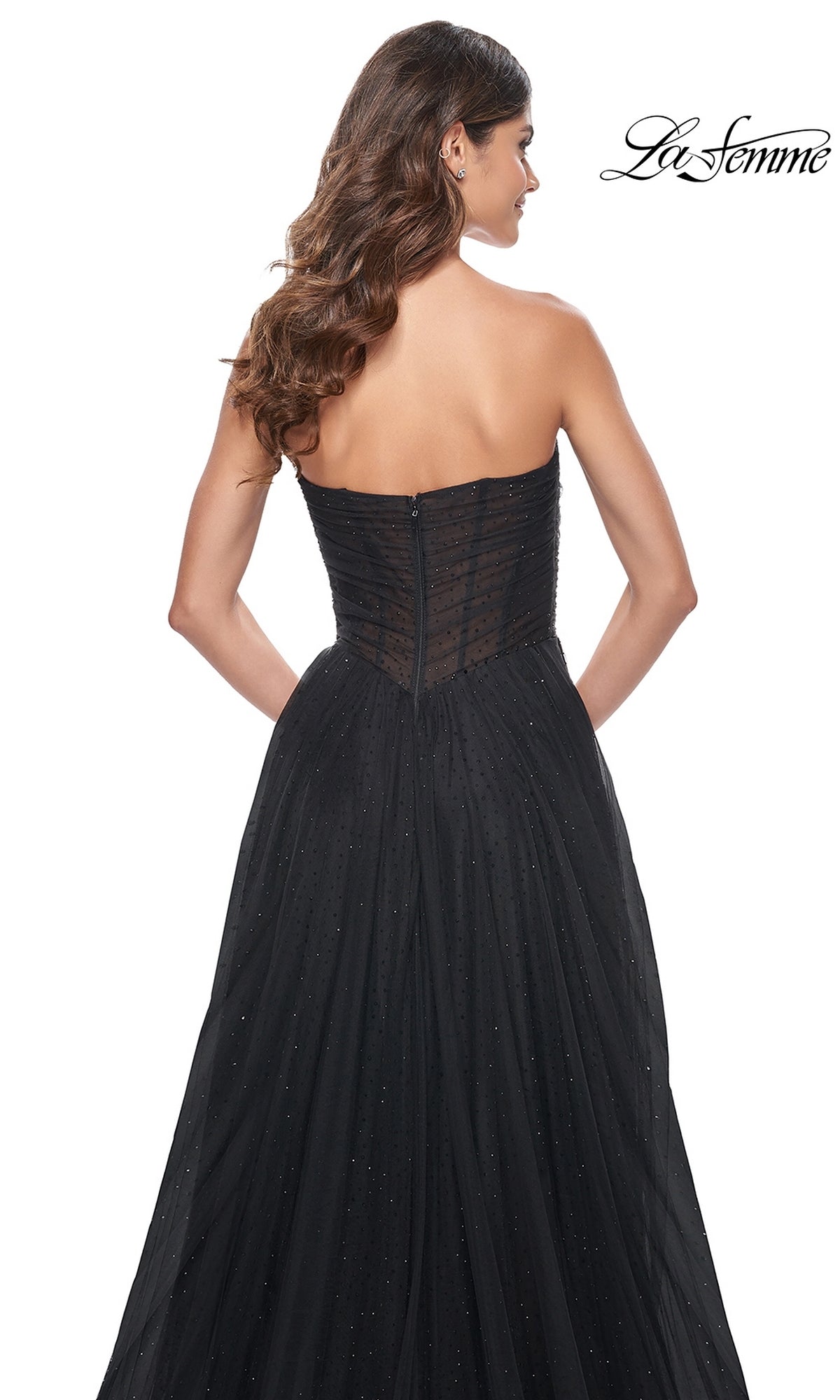 A woman with long wavy hair models the La Femme Long Black Strapless Prom Ball Gown 32029, featuring a sheer, fitted bodice and flowing skirt, shown from the back.